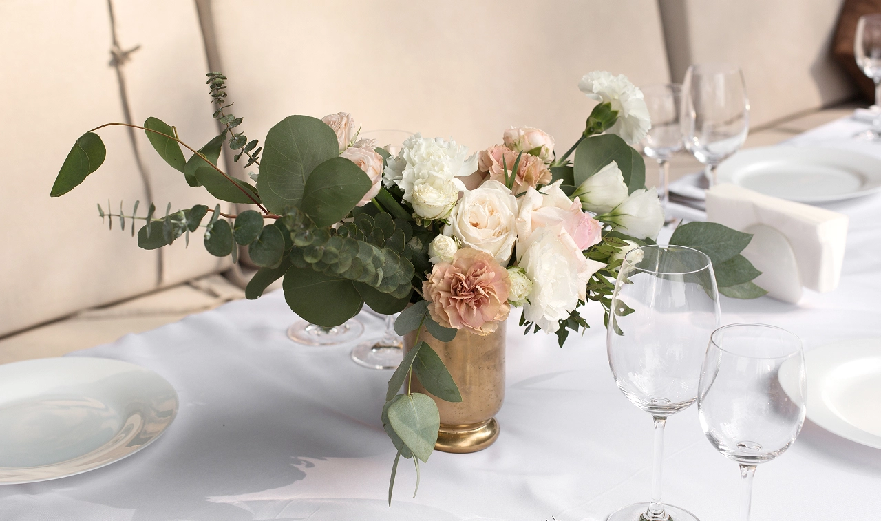 Elegant floral centerpiece with white and soft pink roses and greenery in a gold vase on a white tablecloth with empty wine glasses and plates.
