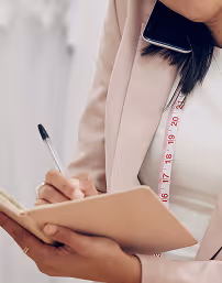 Persona escribiendo en una libreta con un teléfono móvil entre el hombro y la oreja, y una cinta métrica colgando del cuello.
