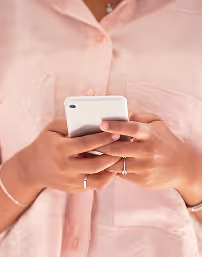 Persona con camisa rosa usando un teléfono celular blanco, enfocada en sus manos.