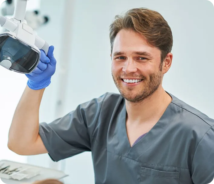 Smiling male dentist in gray scrubs holding dental light in a bright clinic.