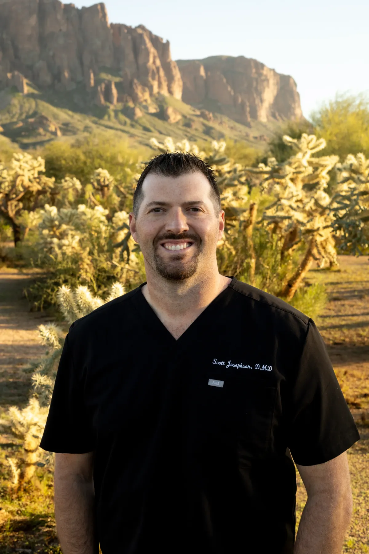 Smiling man in black scrubs standing outdoors with desert plants and rocky mountains in the background.