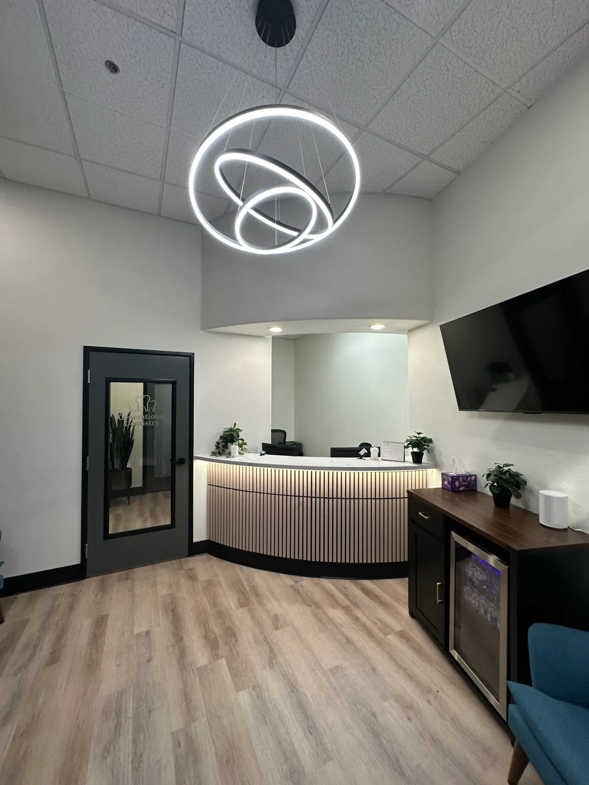 Modern reception area with circular LED chandelier, curved desk, TV, plants, and hardwood floor.