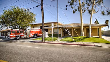 Two red fire trucks parked outside a single-story fire station with green lawn and trees.