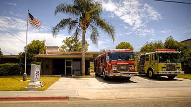 Two fire trucks parked outside a fire station with palm trees and an American flag in front.
