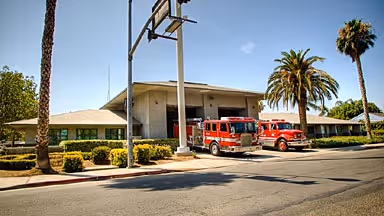 Two red fire trucks parked outside a beige fire station building with palm trees around.