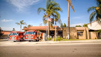 Two red fire trucks parked outside a fire station with palm trees and an American flag in front.