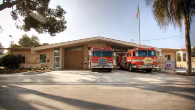 Two red fire trucks parked in front of a single-story fire station building with a flagpole and trees nearby.