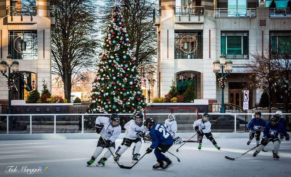 Children playing ice hockey on an outdoor rink decorated with a large Christmas tree in the background.