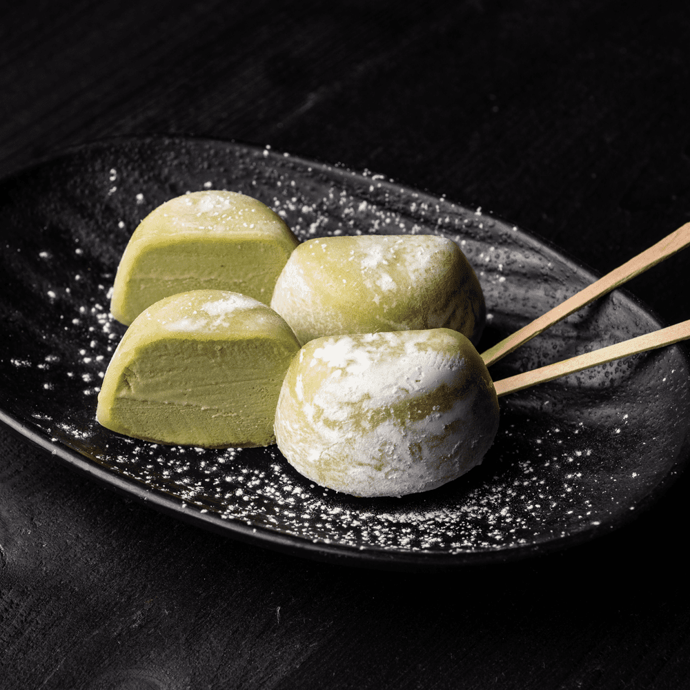 Green matcha mochi ice cream balls on a black plate dusted with powdered sugar, two skewered with wooden sticks.