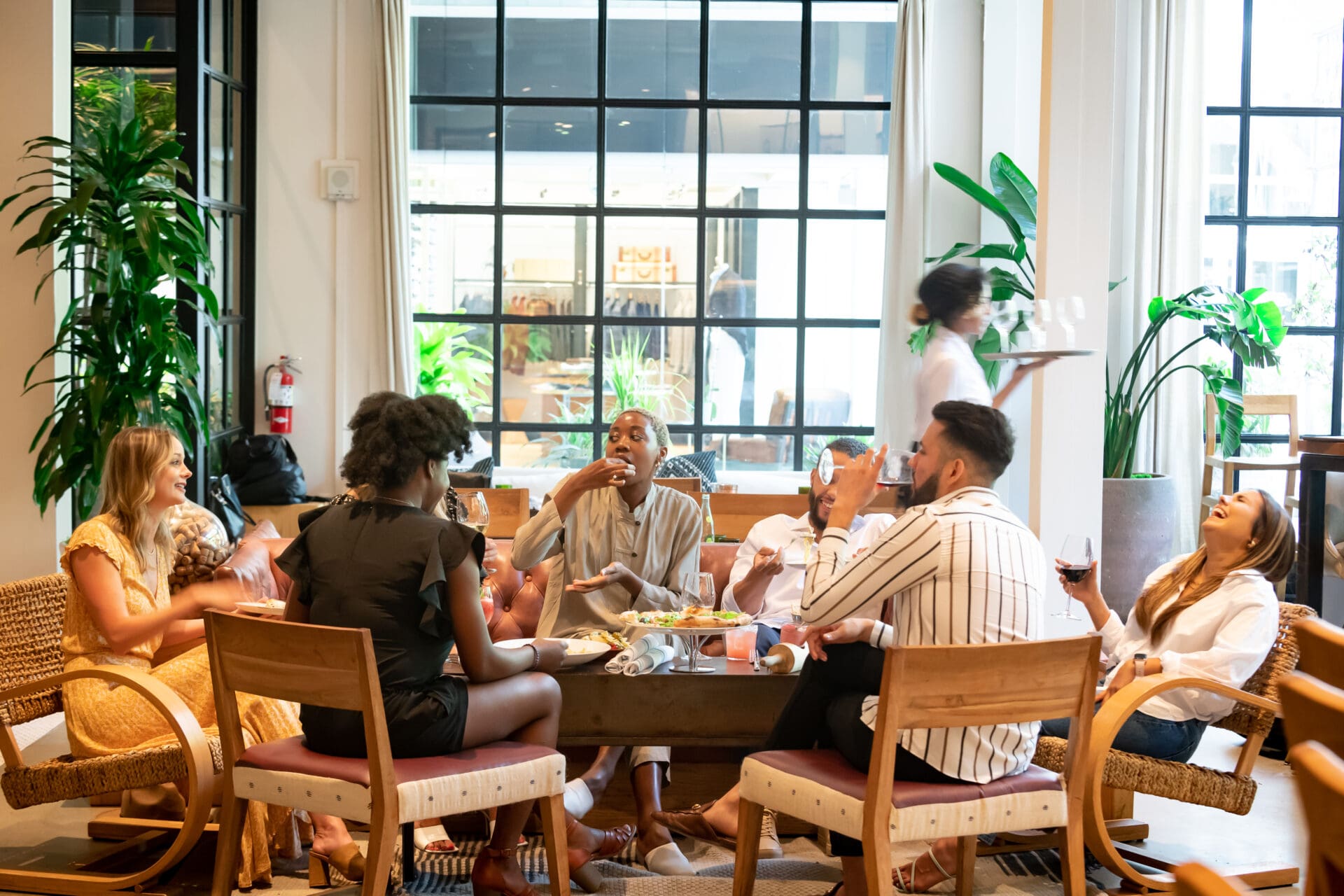 Group of six diverse people sitting around a table in a bright dining area, eating, drinking wine, and laughing while a server walks by.