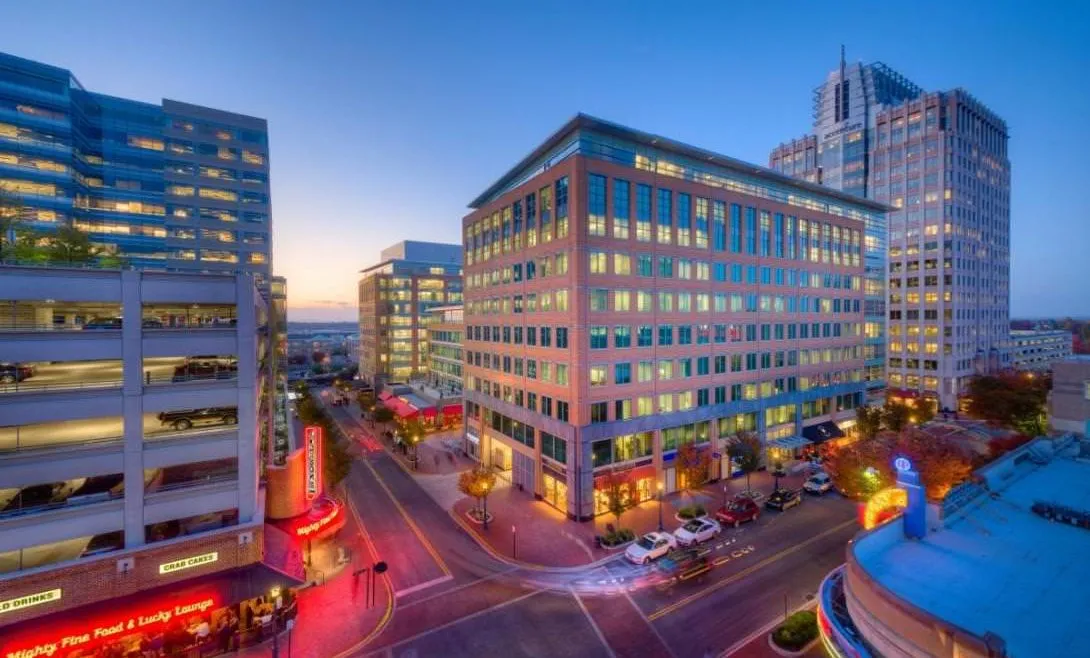 Evening view of a city intersection with illuminated modern office buildings and parked cars.