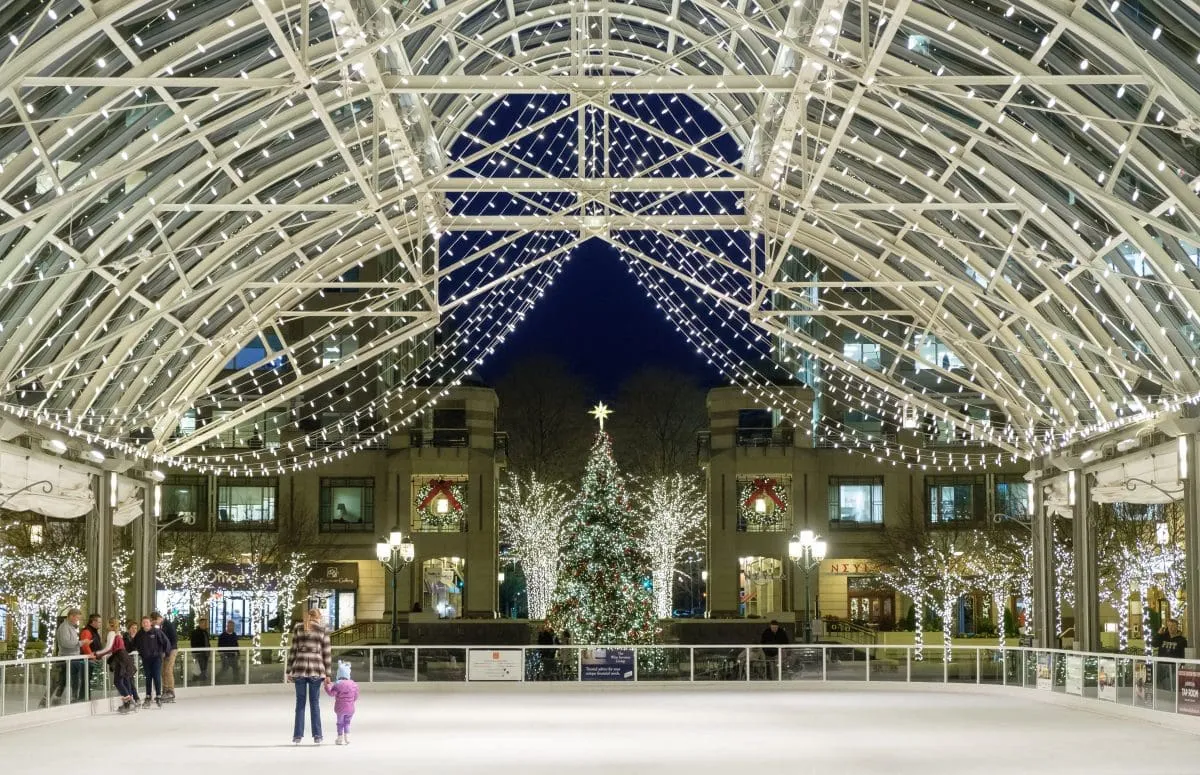 People ice skating inside a lit glass pavilion decorated with string lights and a large Christmas tree in the background at night.