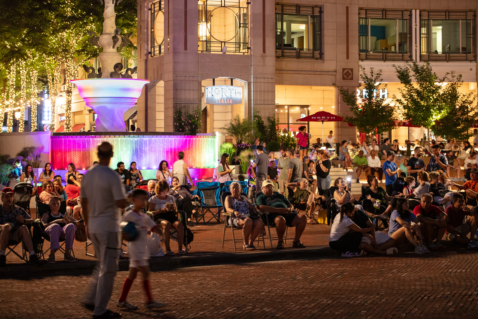 Crowd of people gathered at night in a plaza with a large illuminated fountain and building with 'North Italia' sign in the background.