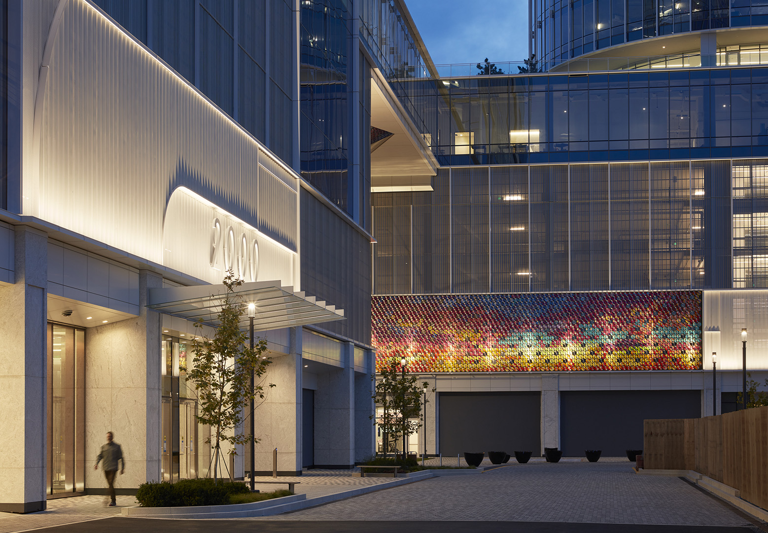 Modern office building at dusk with illuminated '2000' sign and colorful mosaic wall art, a person walking near the entrance.