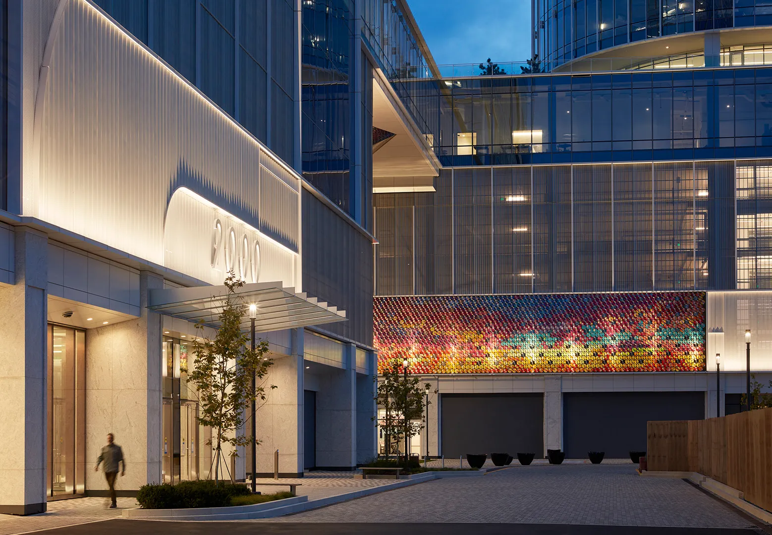 Modern office building at dusk with illuminated '2000' sign and colorful mosaic wall art, a person walking near the entrance.