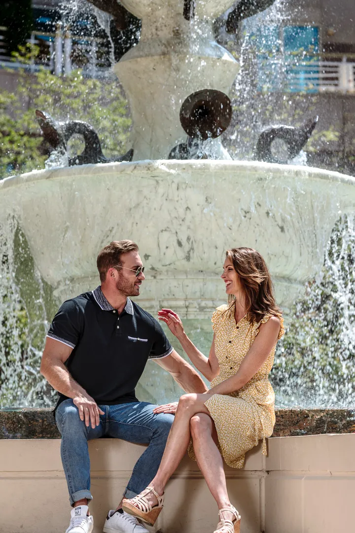 Man and woman sitting and smiling while talking in front of a large stone water fountain outdoors.