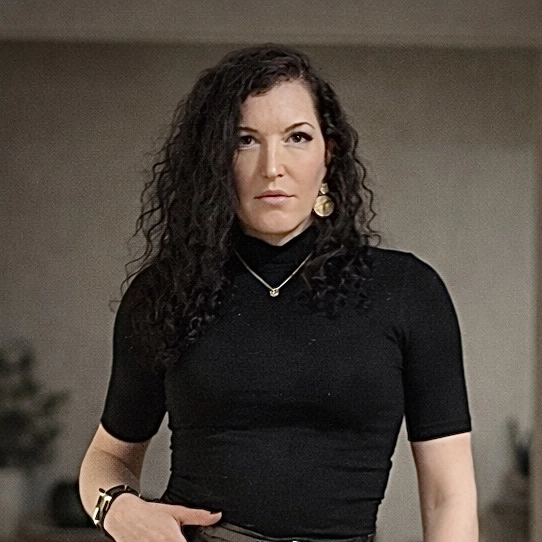 Portrait d'Olivia Cefai en 2026. Femme aux cheveux bouclés, portant un haut noir à manches courtes et des boucles d'oreilles dorées, regardant sérieusement devant elle.