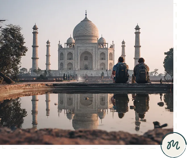 Two travelers with backpacks sitting by a reflecting pool facing the Taj Mahal at sunset.