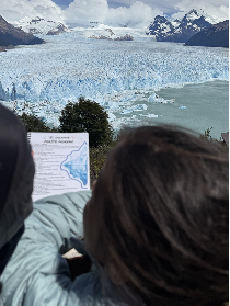 People viewing a glacier with an informational sign about the glacier in the foreground and snowy mountains in the background.