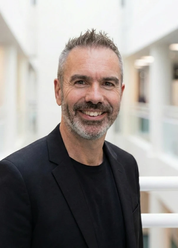 Smiling middle-aged man with short graying hair and beard wearing a black blazer and shirt in a bright indoor setting.