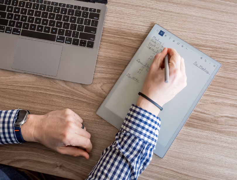 Person writing notes with a stylus on a digital tablet beside a laptop keyboard on a wooden desk.
