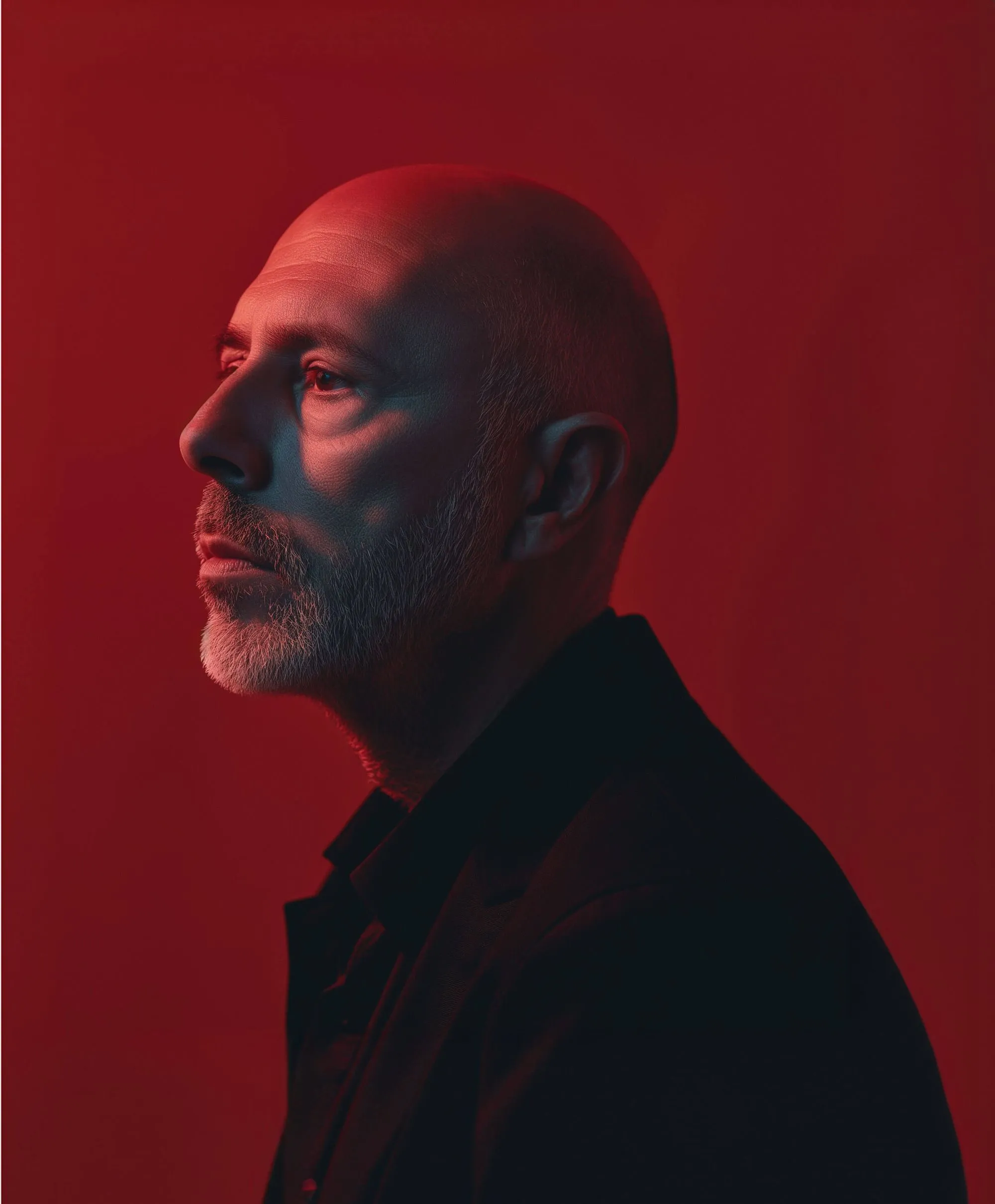 Portrait d'un homme chauve avec barbe grise sur fond rouge, éclairé latéralement en bleu et rouge.