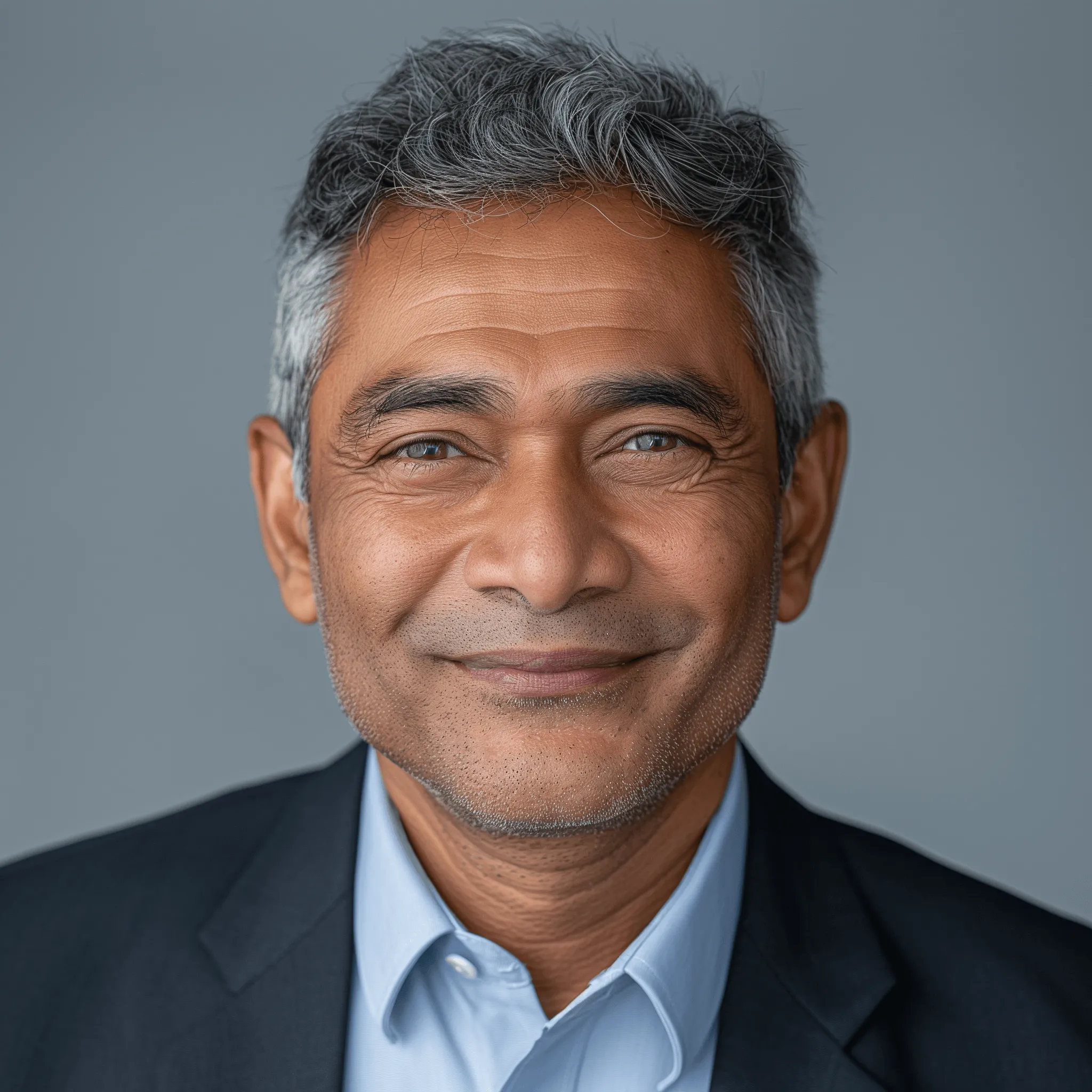Portrait d'un homme mûr souriant avec cheveux poivre et sel, portant une chemise bleue et un blazer noir.