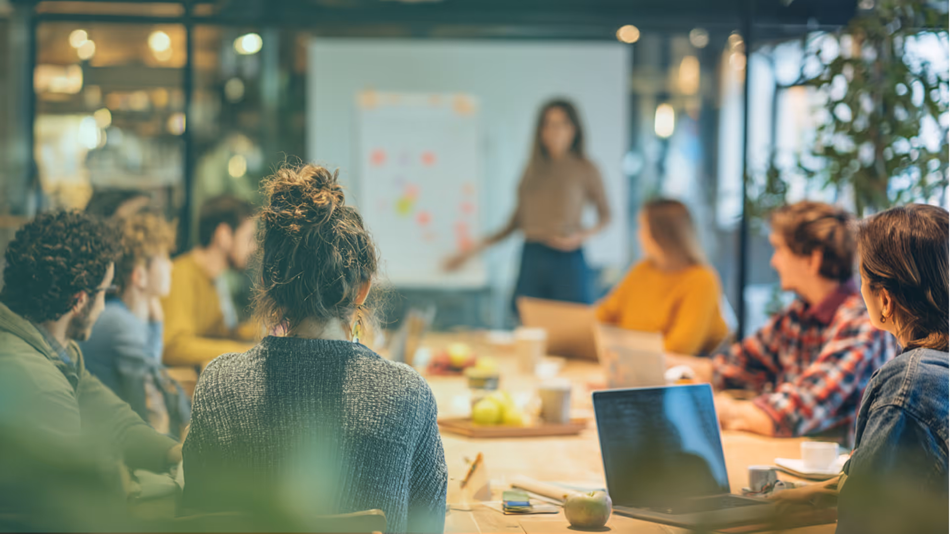 Un groupe de personnes assises à une table, écoutant une femme qui présente devant un tableau. Session de formation et d'échange entre professionnels.