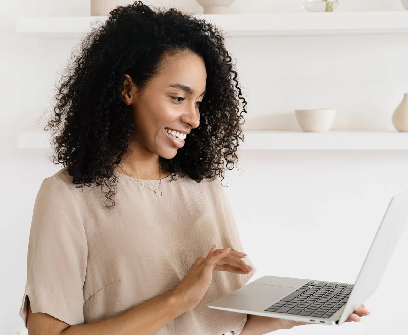 Smiling woman with curly hair using a laptop in a bright, minimalist room.