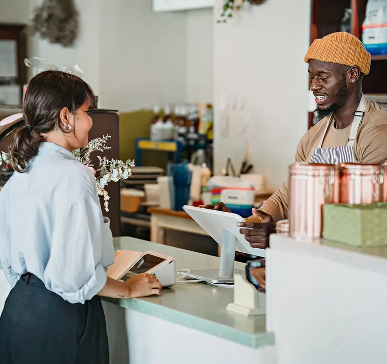 Barista wearing a striped apron and orange beanie assisting a female customer at a cafe counter with a tablet register.