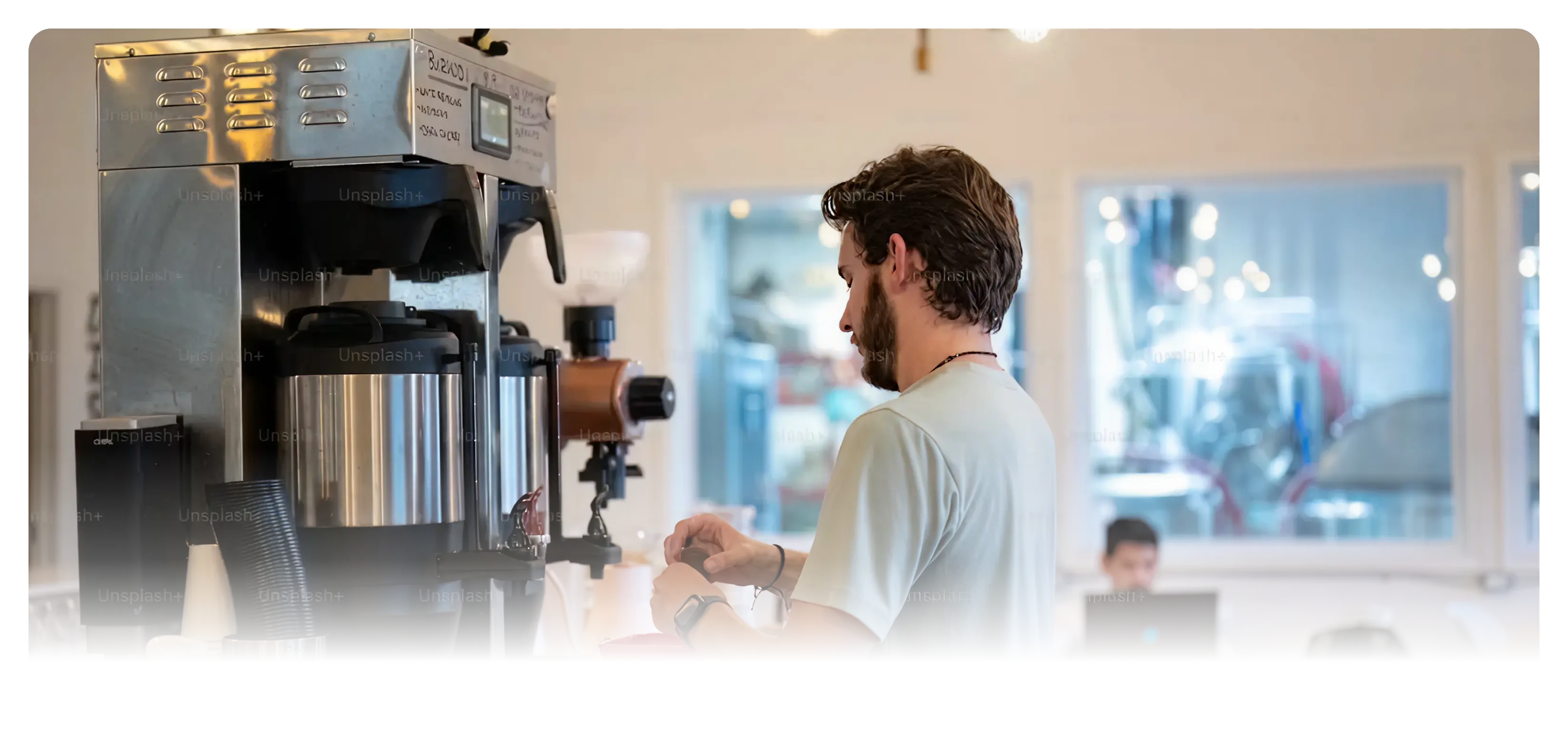 Man in a light gray shirt preparing coffee near large coffee dispensers in a bright room with another person working on a laptop in the background.