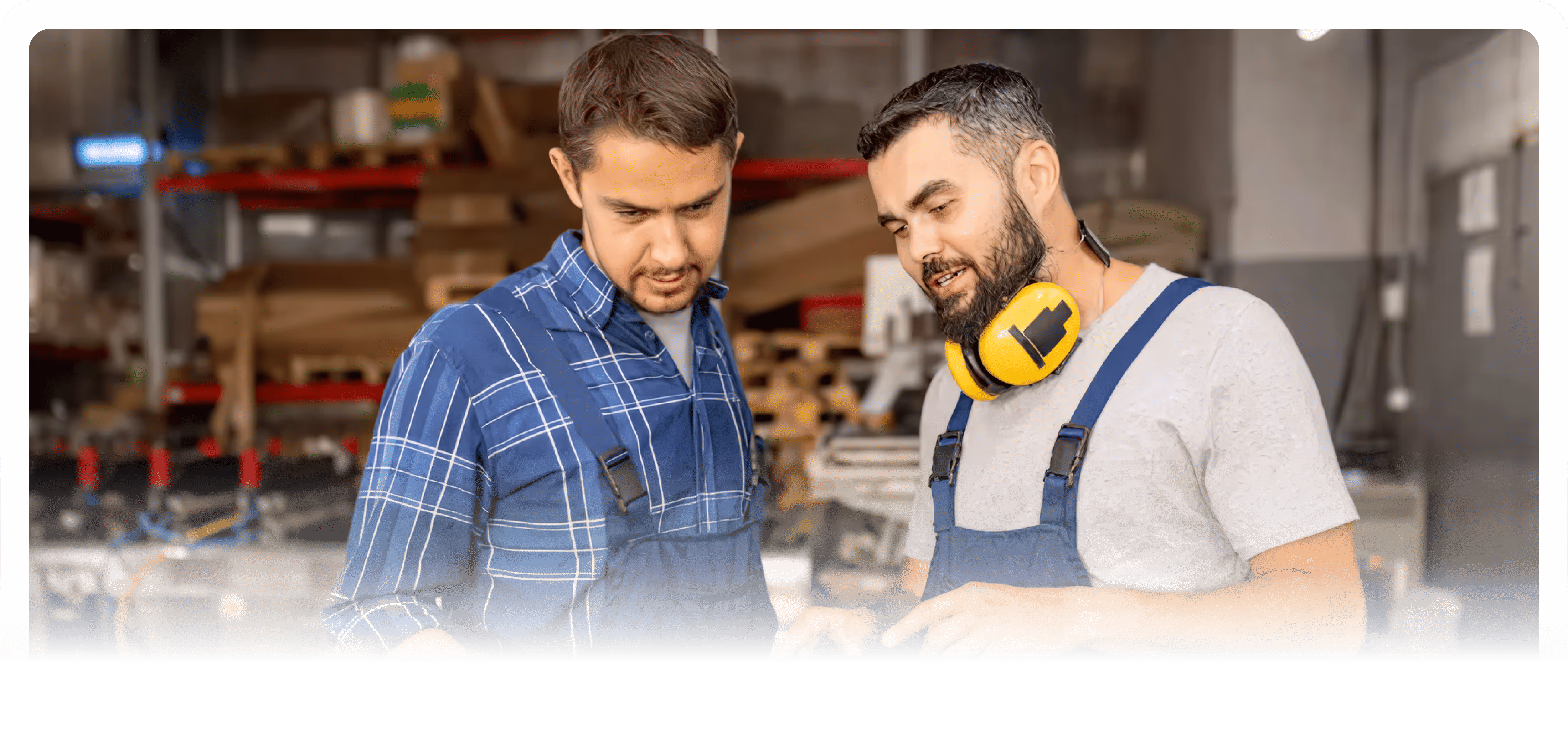 Two men in work overalls discussing plans in a workshop, one wearing yellow ear protection around his neck.