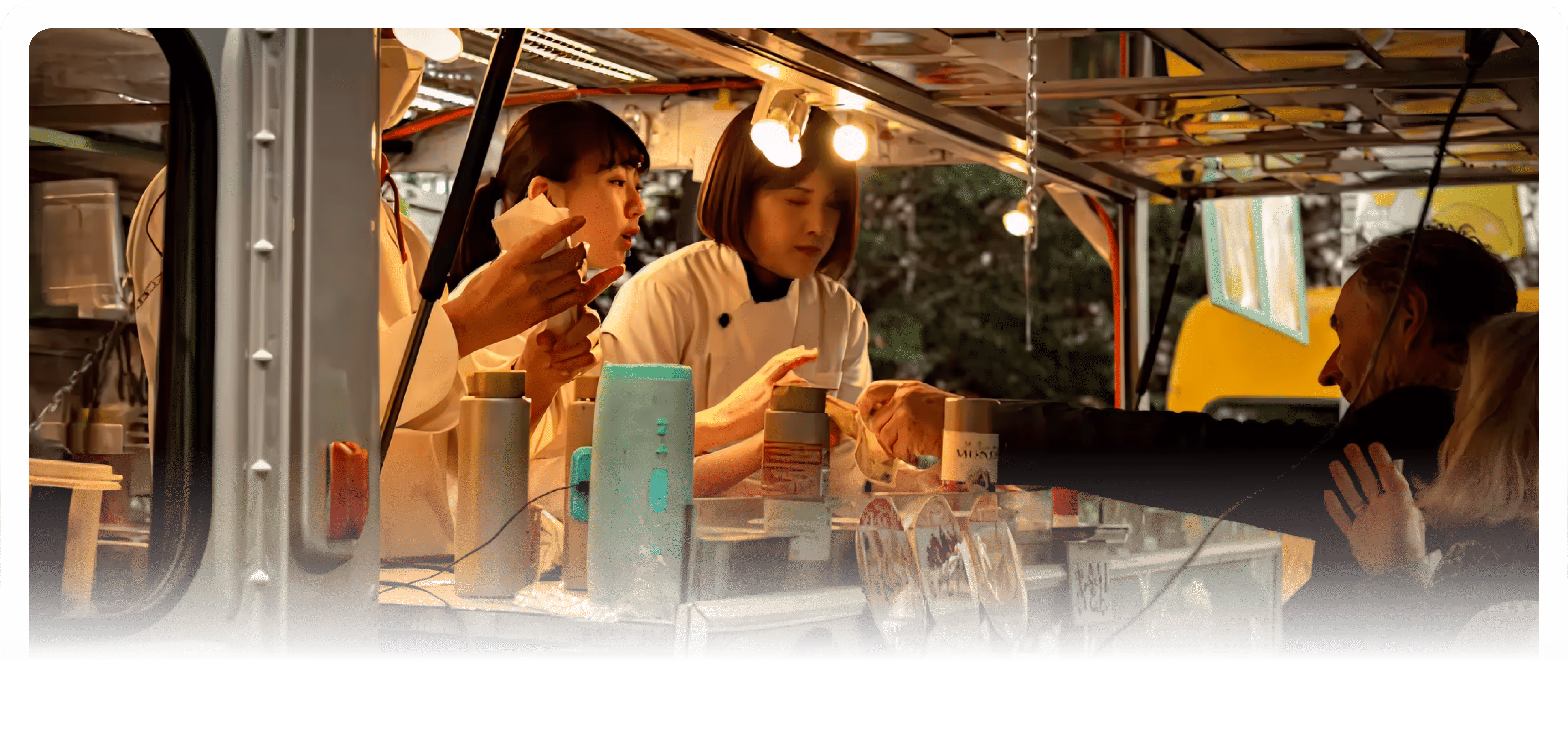 Two women in white uniforms serving food to customers at an outdoor food truck with illuminated menu signs.