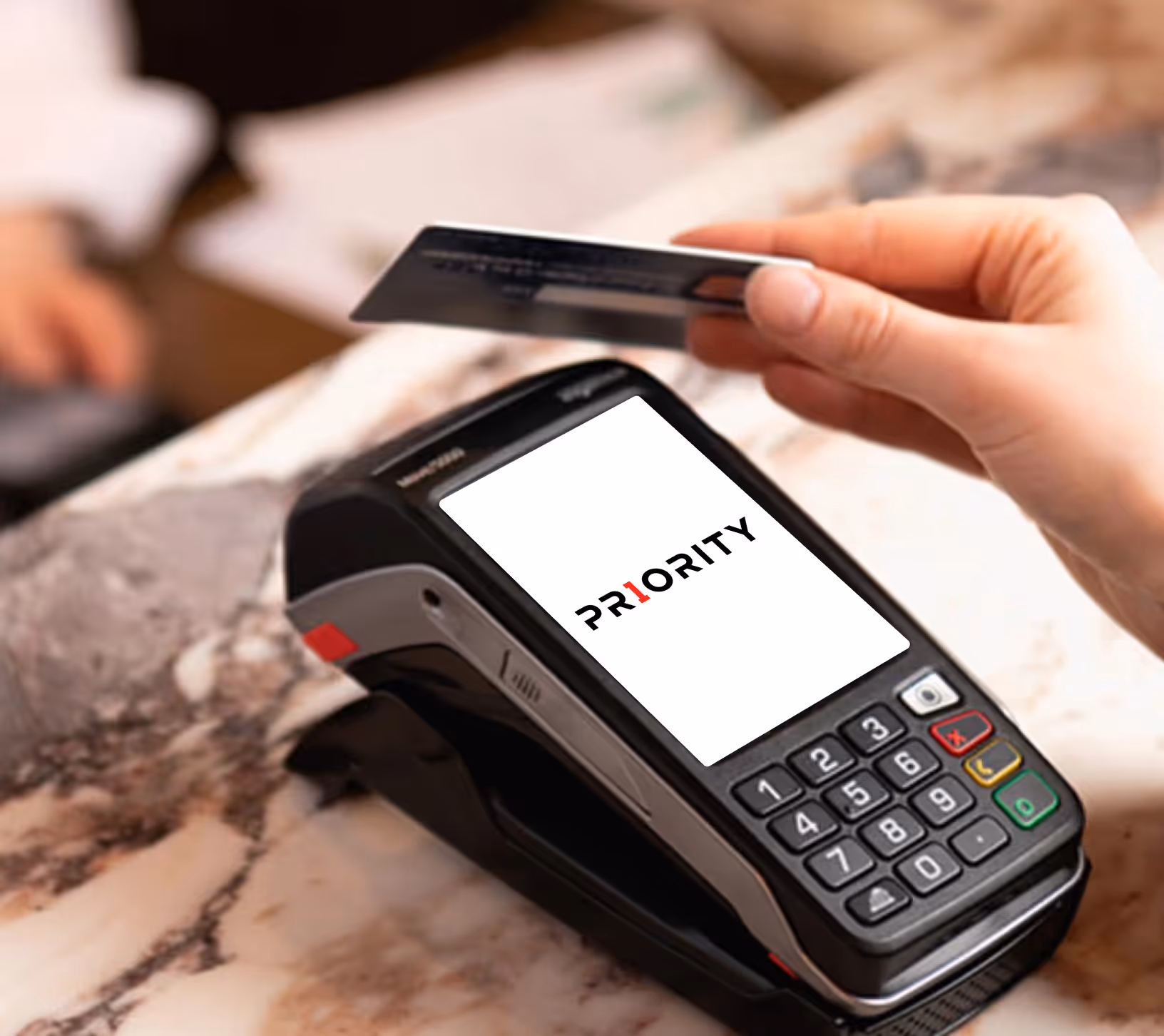 Hand holding a credit card near a payment terminal displaying the word PRIORITY on the screen.