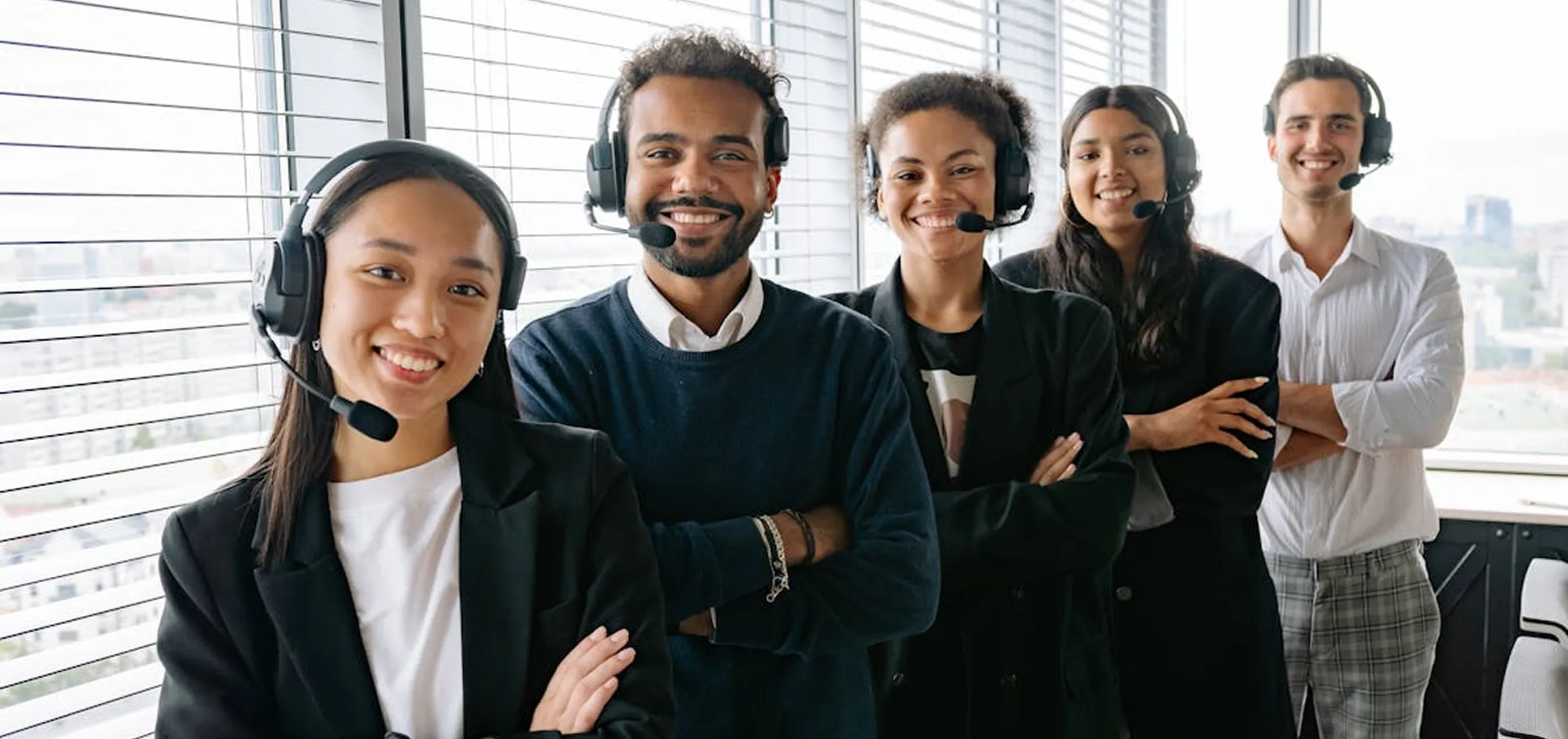 Diverse customer service team wearing headsets and smiling inside an office with window blinds.