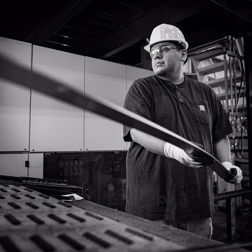 Worker wearing a hard hat and safety glasses holding a metal sheet in an industrial setting.