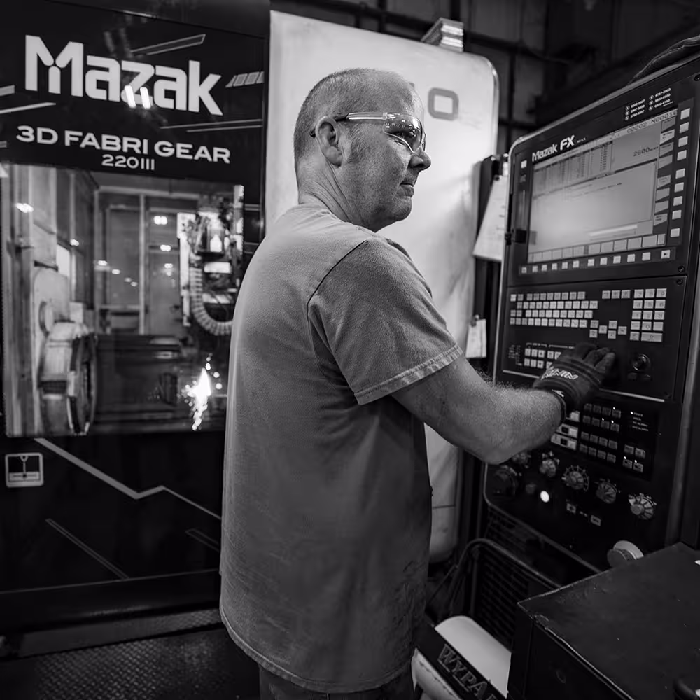 Worker wearing safety glasses and gloves operating a Mazak 3D Fabri Gear 220III CNC machine.
