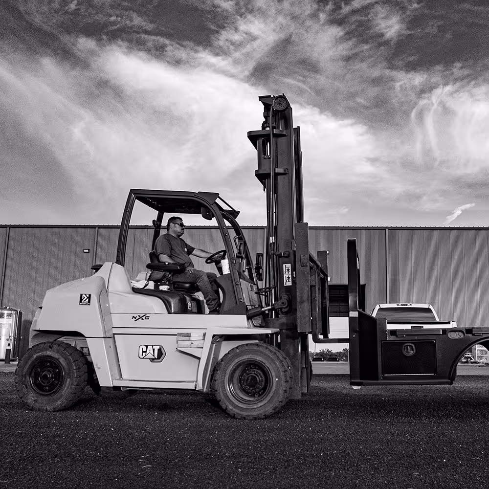 Man operating a CAT NXG forklift outdoors on gravel with a large building and cloudy sky in the background.