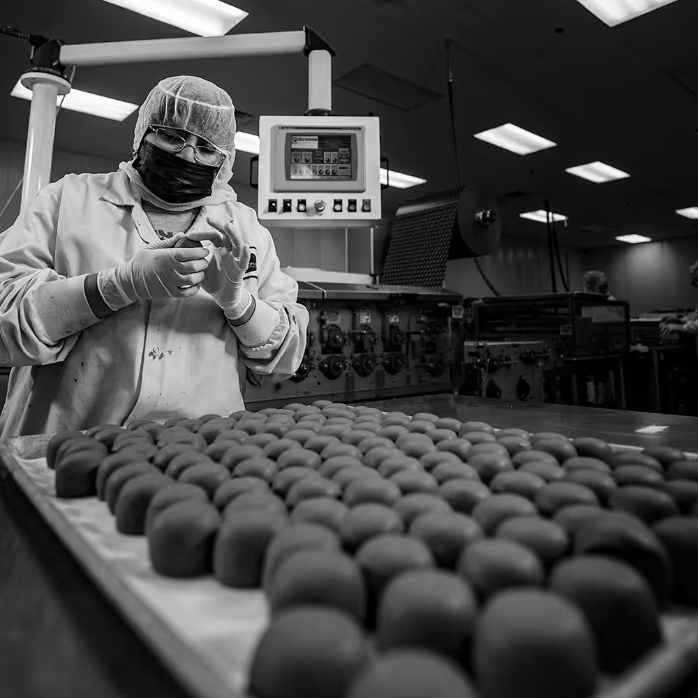 Food production worker in protective gear inspecting small round food items on a tray in a factory setting.
