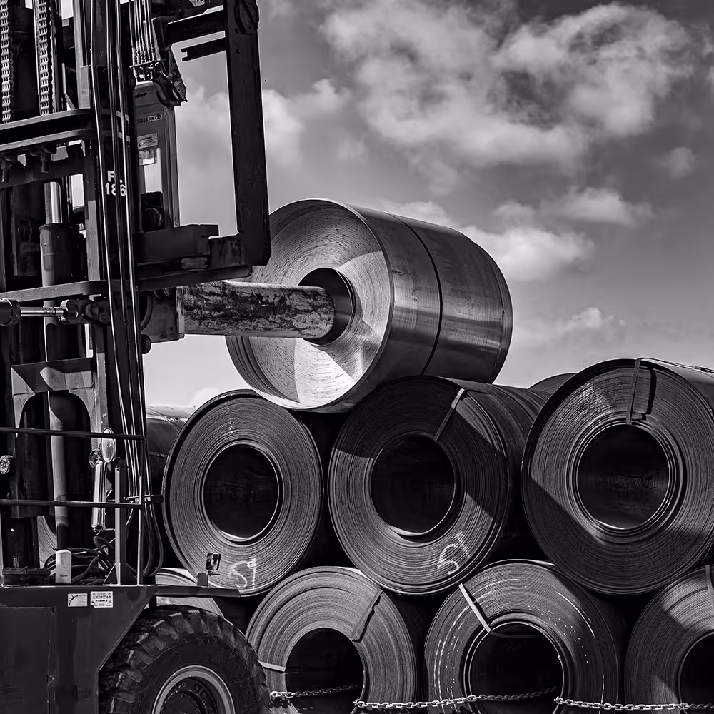 Forklift lifting a large metal coil with stacked metal coils in the background under a partly cloudy sky.