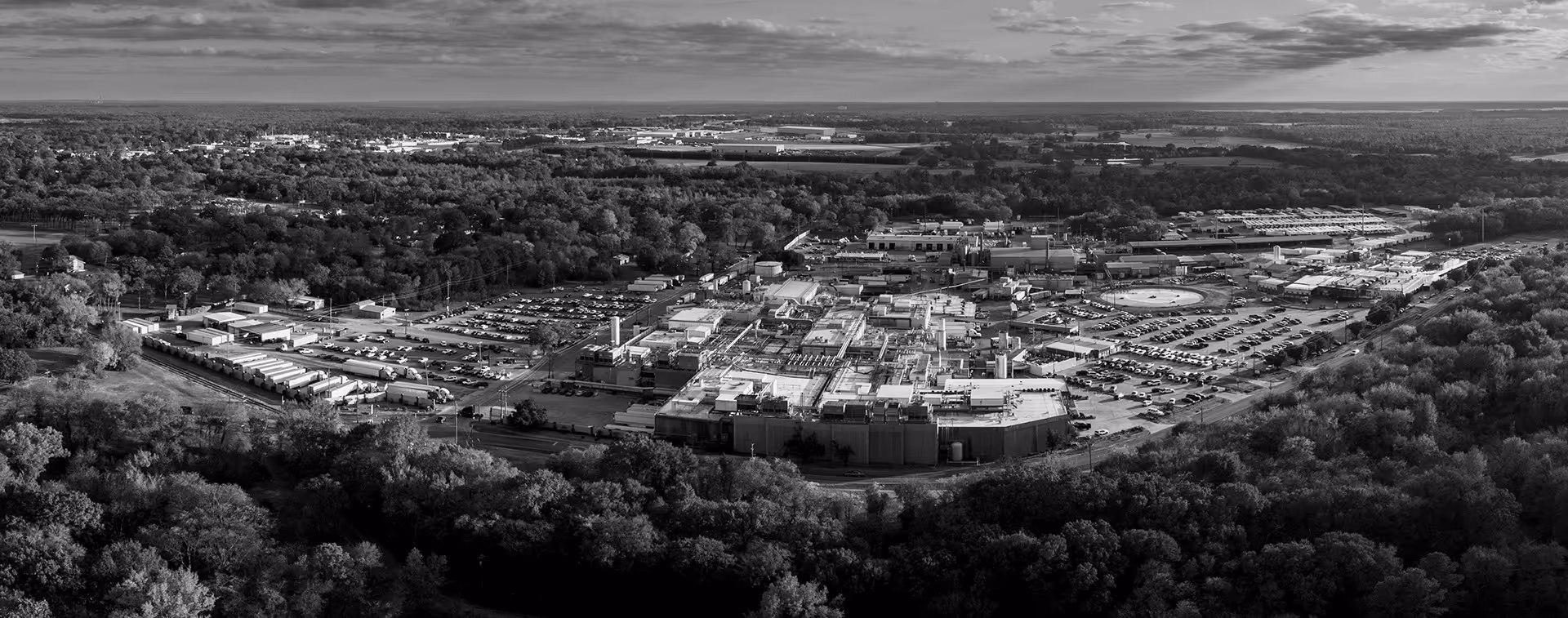 Aerial view of an industrial complex surrounded by dense trees and parking lots under a partly cloudy sky.