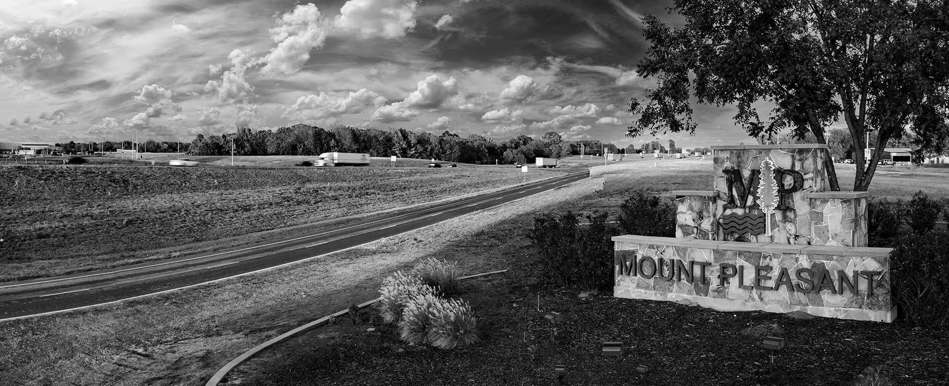 Stone sign with 'Mount Pleasant' text and tree logo beside a highway under a cloudy sky.