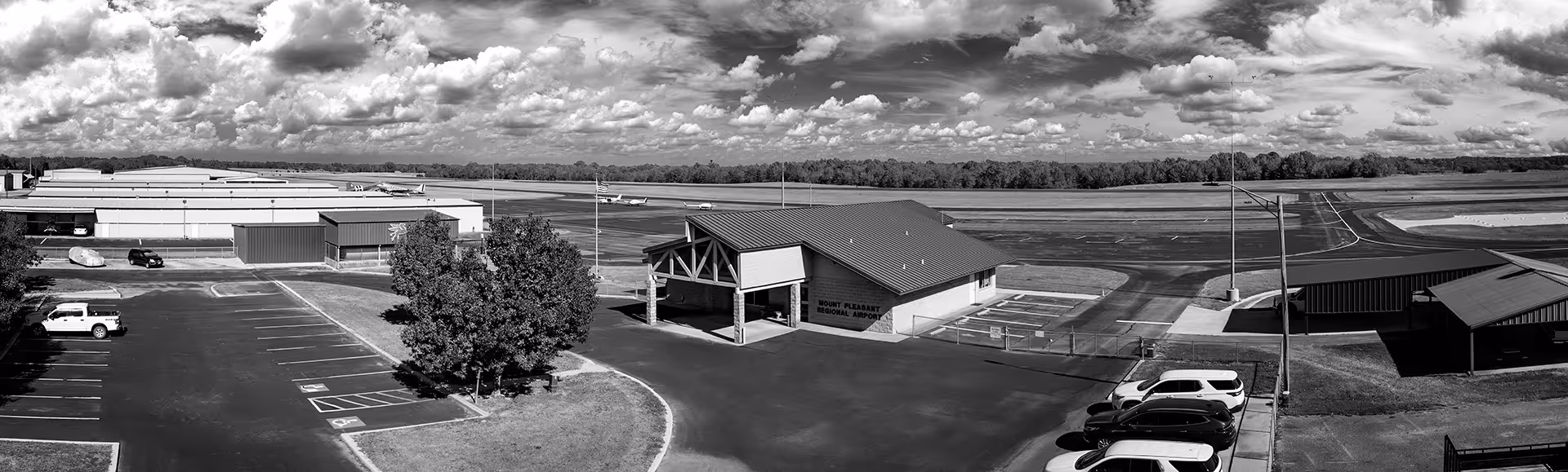 Mount Pleasant Regional Airport terminal building with adjacent parking lots and planes on the runway under a partly cloudy sky.