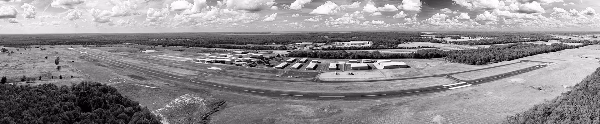 Aerial panoramic view of an airport runway surrounded by hangars, several parked airplanes, and grassy fields under a partly cloudy sky.