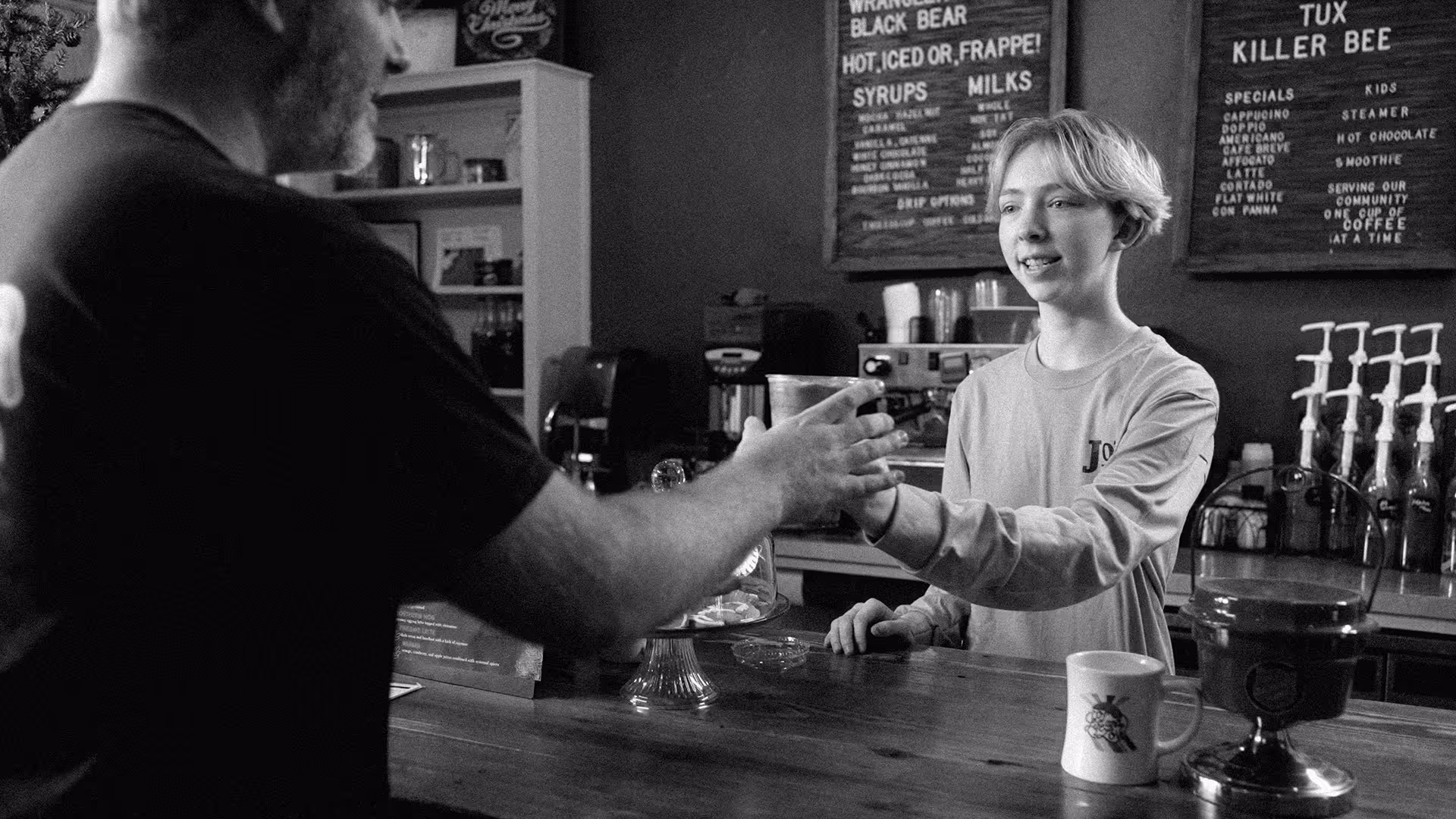 Young barista handing a coffee cup to a customer at a coffee shop counter.
