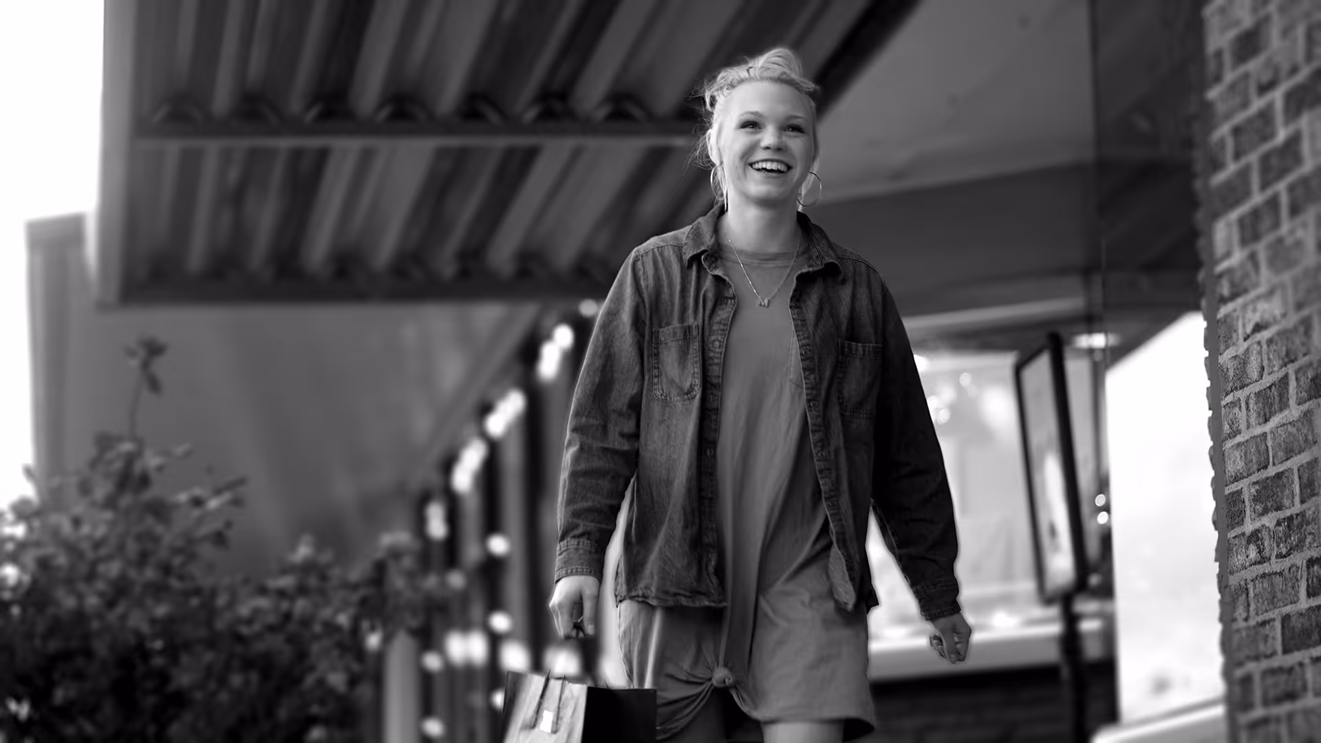Smiling young woman carrying a shopping bag walking outdoors near a storefront.