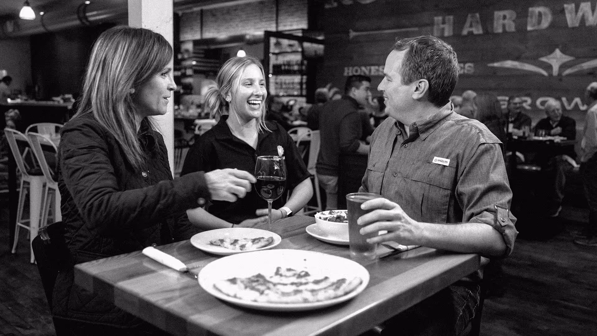 Three people sitting at a wooden table in a restaurant, enjoying pizza, wine, and a drink, and engaging in lively conversation.