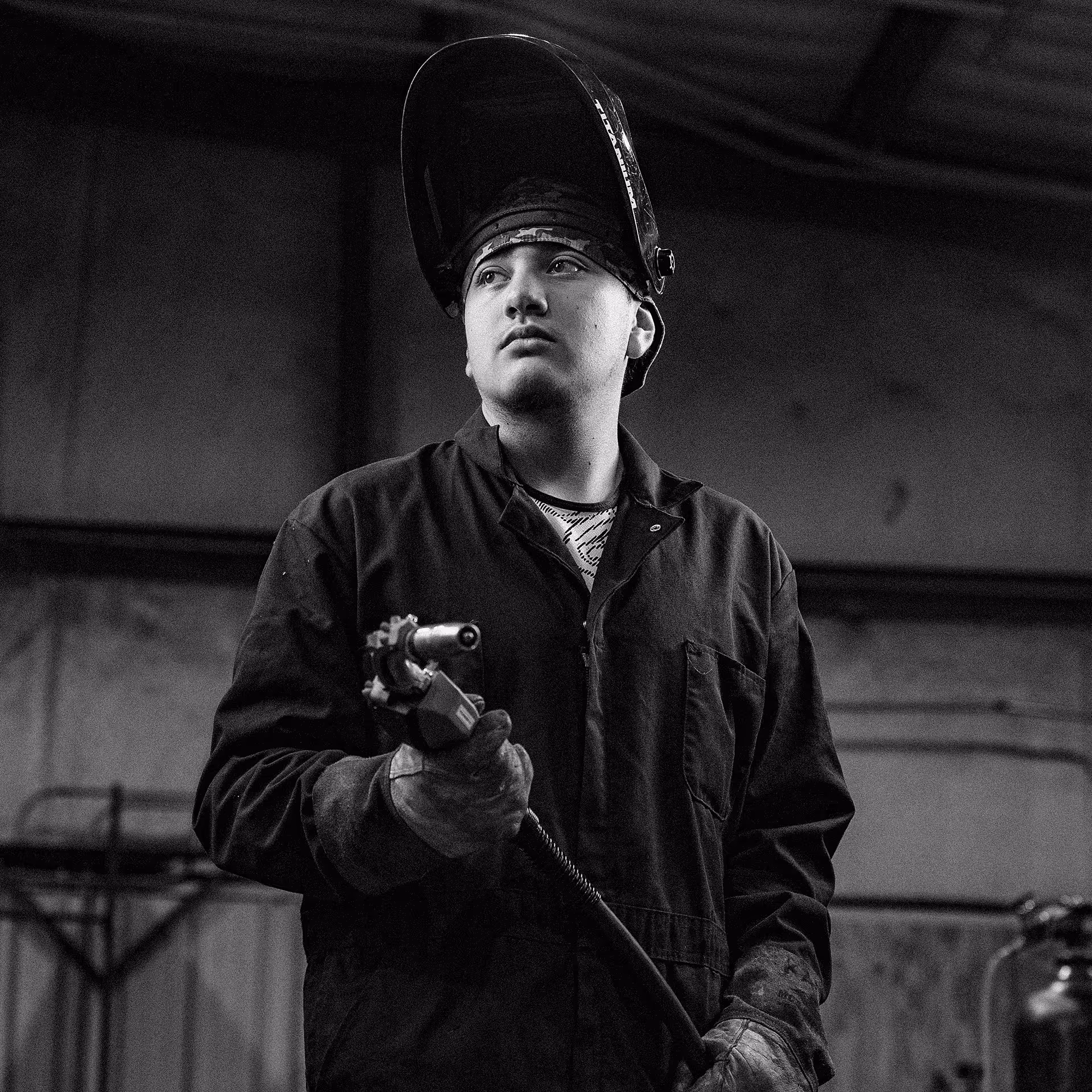 Young welder wearing protective gear holding welding equipment in an industrial workshop.