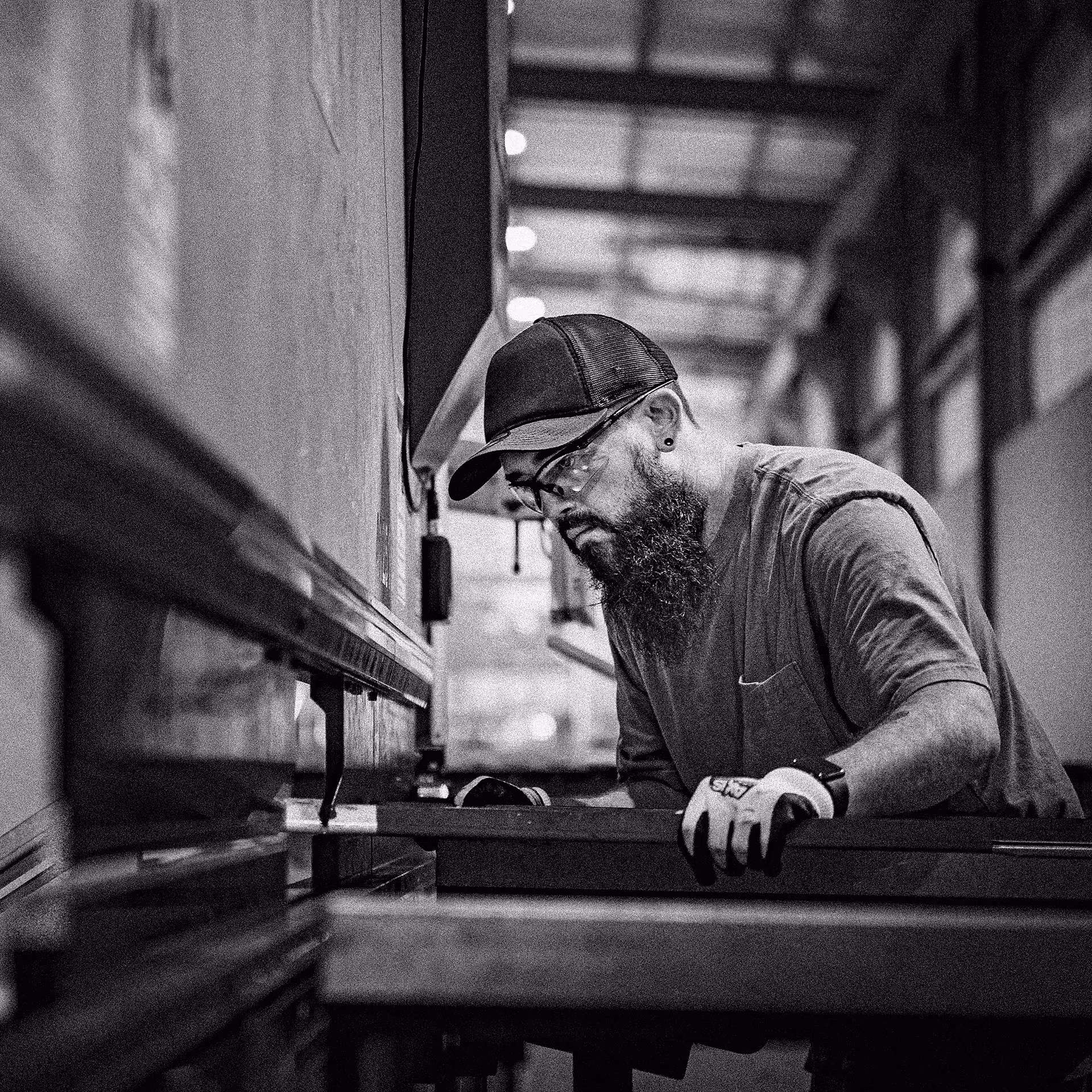 Bearded man wearing safety glasses, a cap, and gloves operating a metalworking machine in a factory.