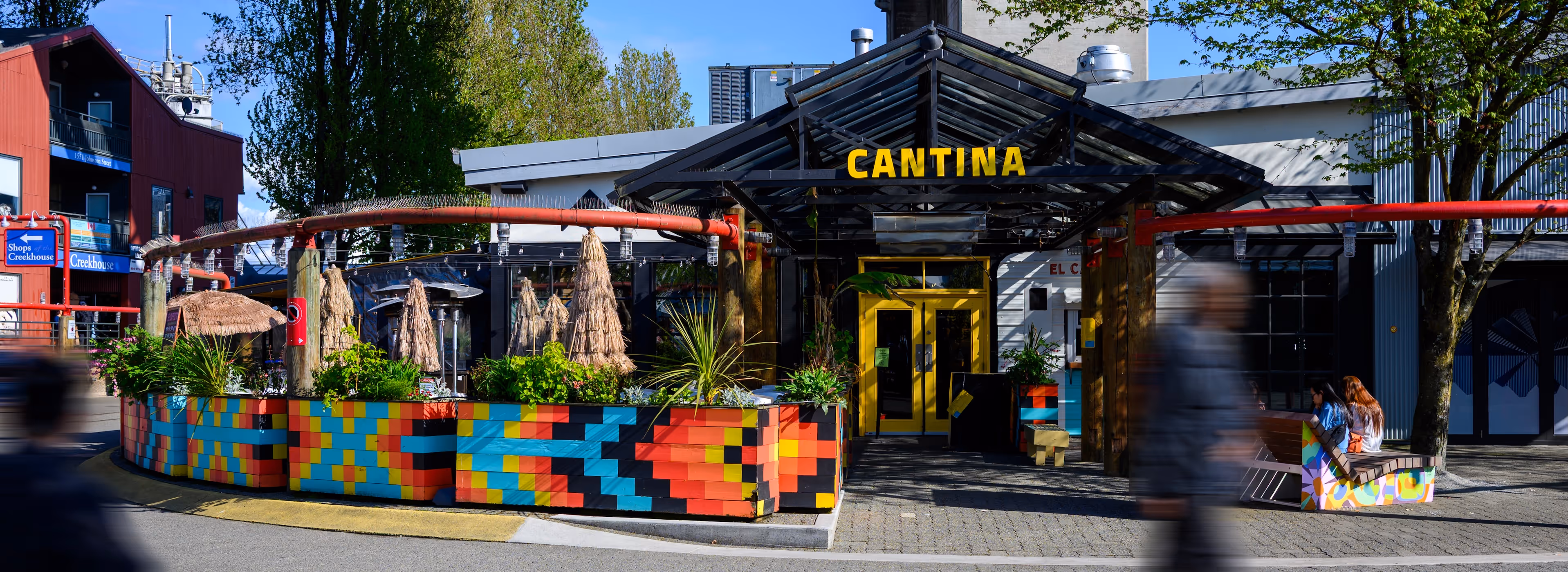 Outdoor entrance to Cantina restaurant with colorful planter boxes, yellow doors, and blurred pedestrians walking by.